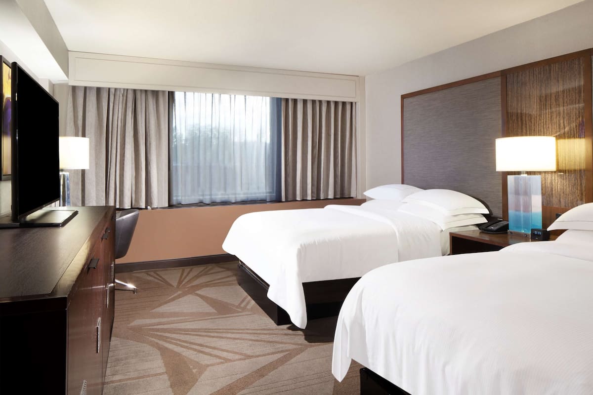 Quarto de hotel com duas camas, uma escrivaninha com televisão, uma luminária e uma janela grande com cortinas.