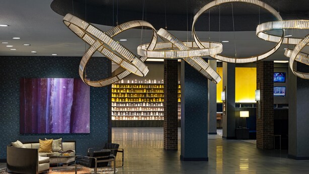 Un hall d’entrée agrémenté d’un lustre moderne, un espace détente avec un canapé et des fauteuils, un grand tableau abstrait et un mur où sont exposées des bouteilles de boissons.