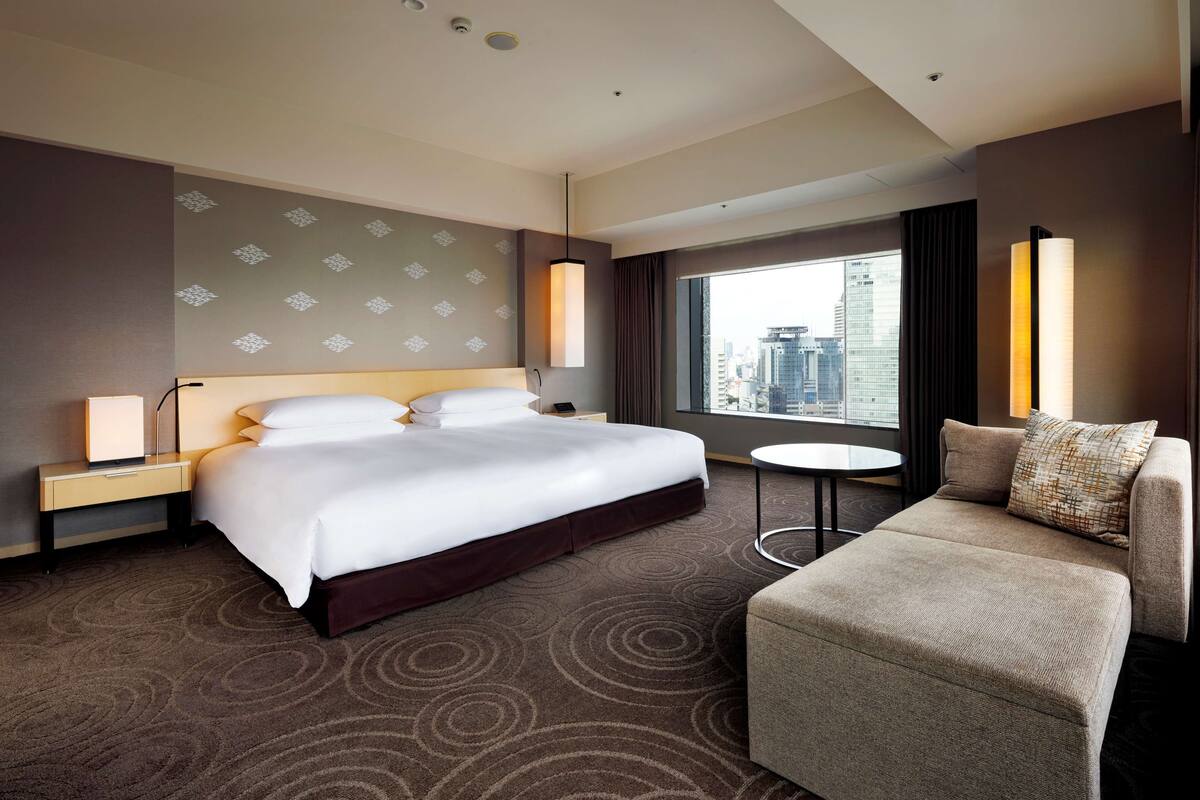 A hotel room with a large bed, a sofa, a small table, and a view of the city through a large window.