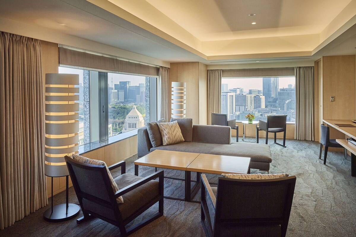 A modern hotel room with a large window offering a city view, a sofa, a dining table, and a desk area.
