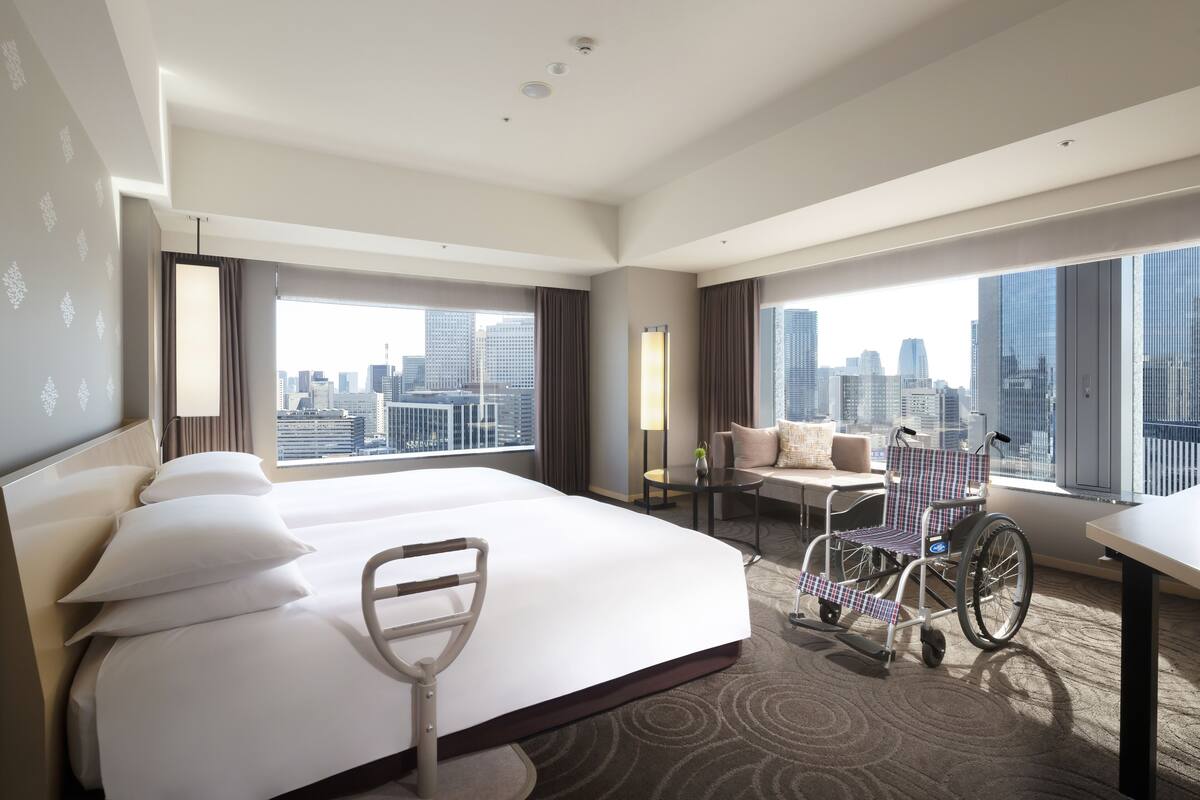 A hotel room with a large bed, wheelchair, city view, and a desk with a chair.
