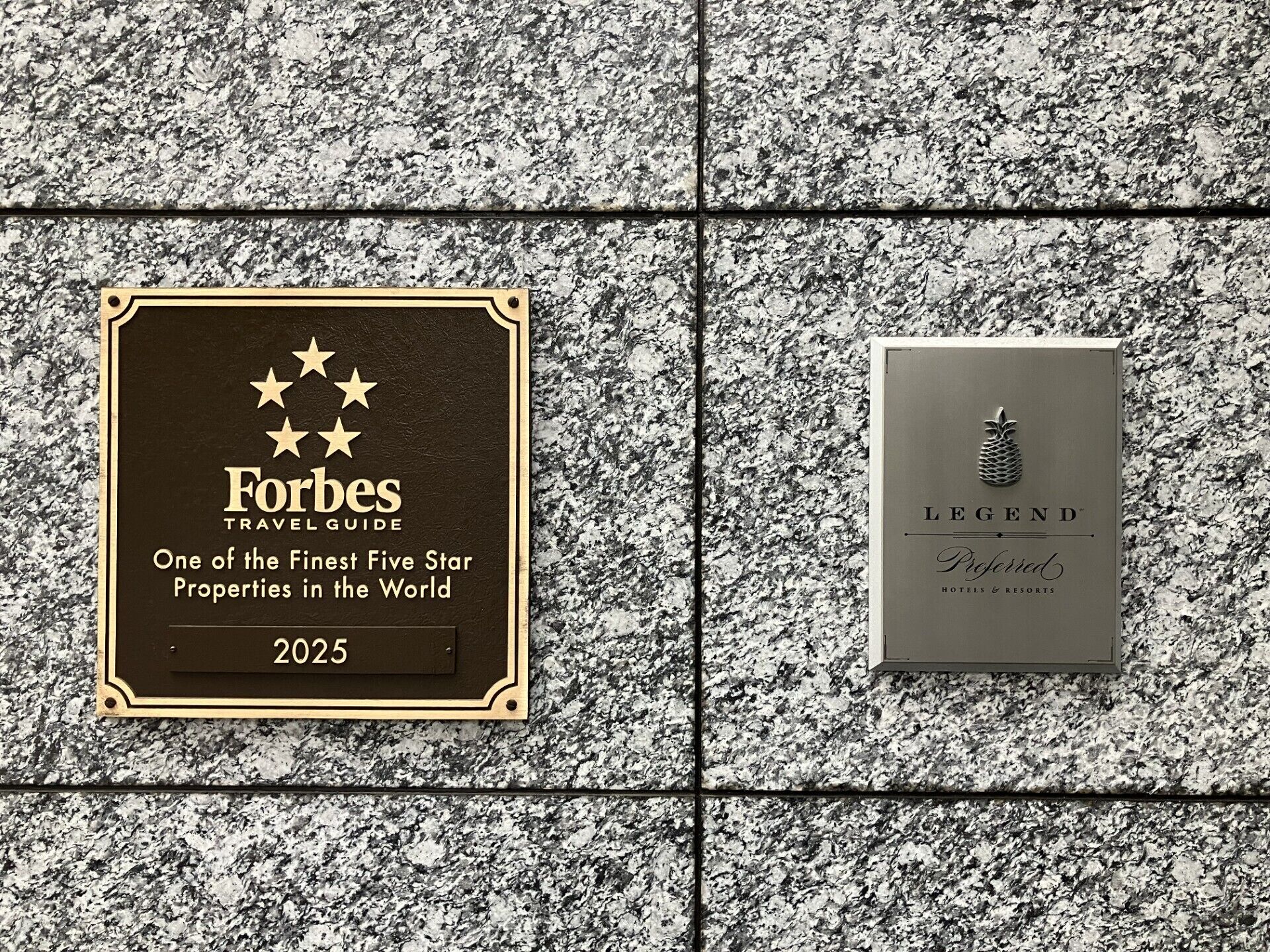 Zwei Schilder an einer Granitwand; eines ist ein Forbes Travel Guide-Schild für 2025, das andere ein Legend Hotels & Resorts-Schild.