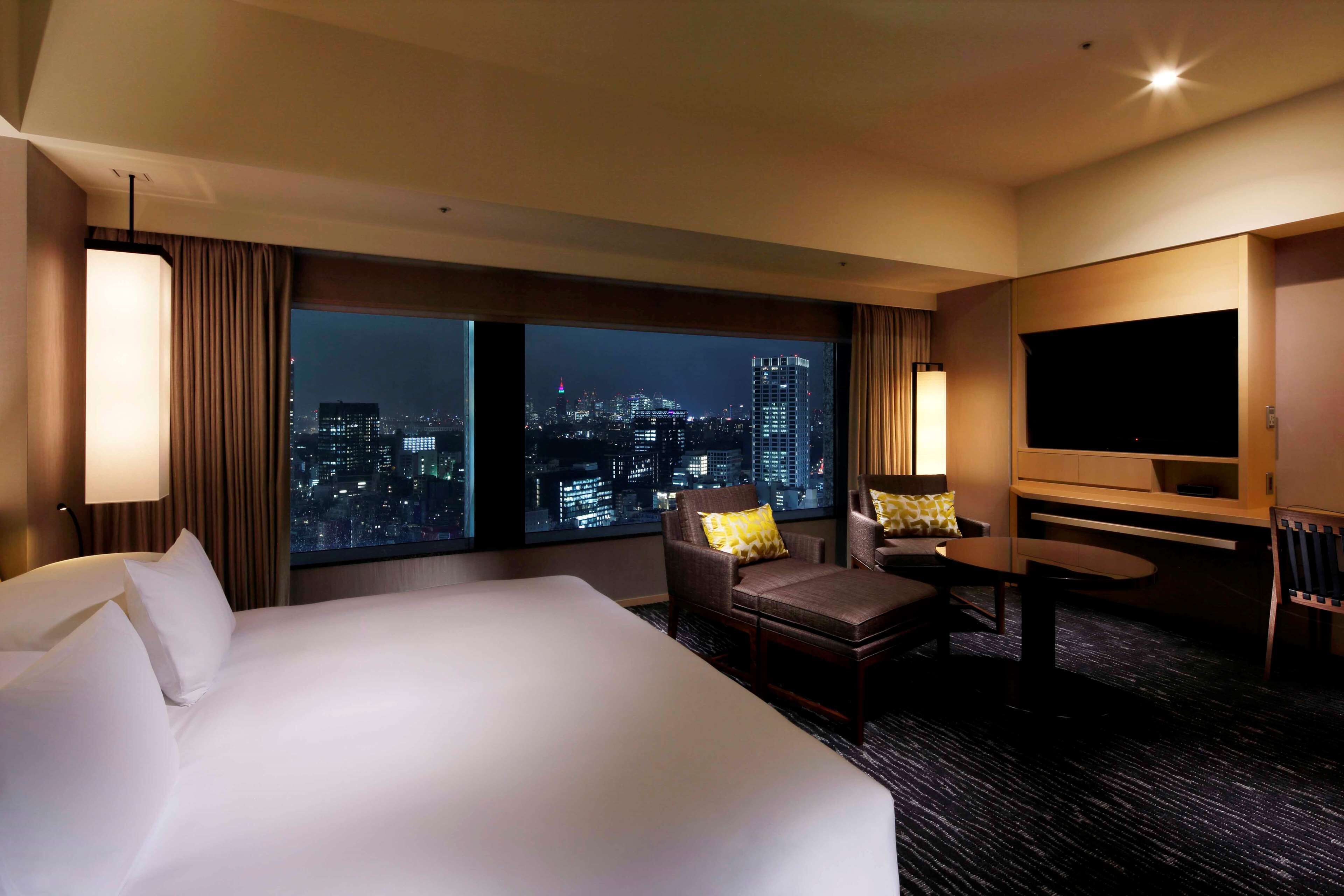 Habitación de hotel con una cama grande, un área de descanso con una mesa y sillas, un televisor de pantalla plana y vistas a la ciudad a través de amplias ventanas.