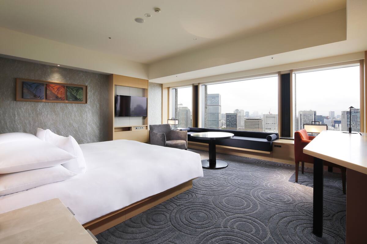 A hotel room with a large bed, a desk, a chair, and a sofa. There is a TV mounted on the wall and a window with a city view.