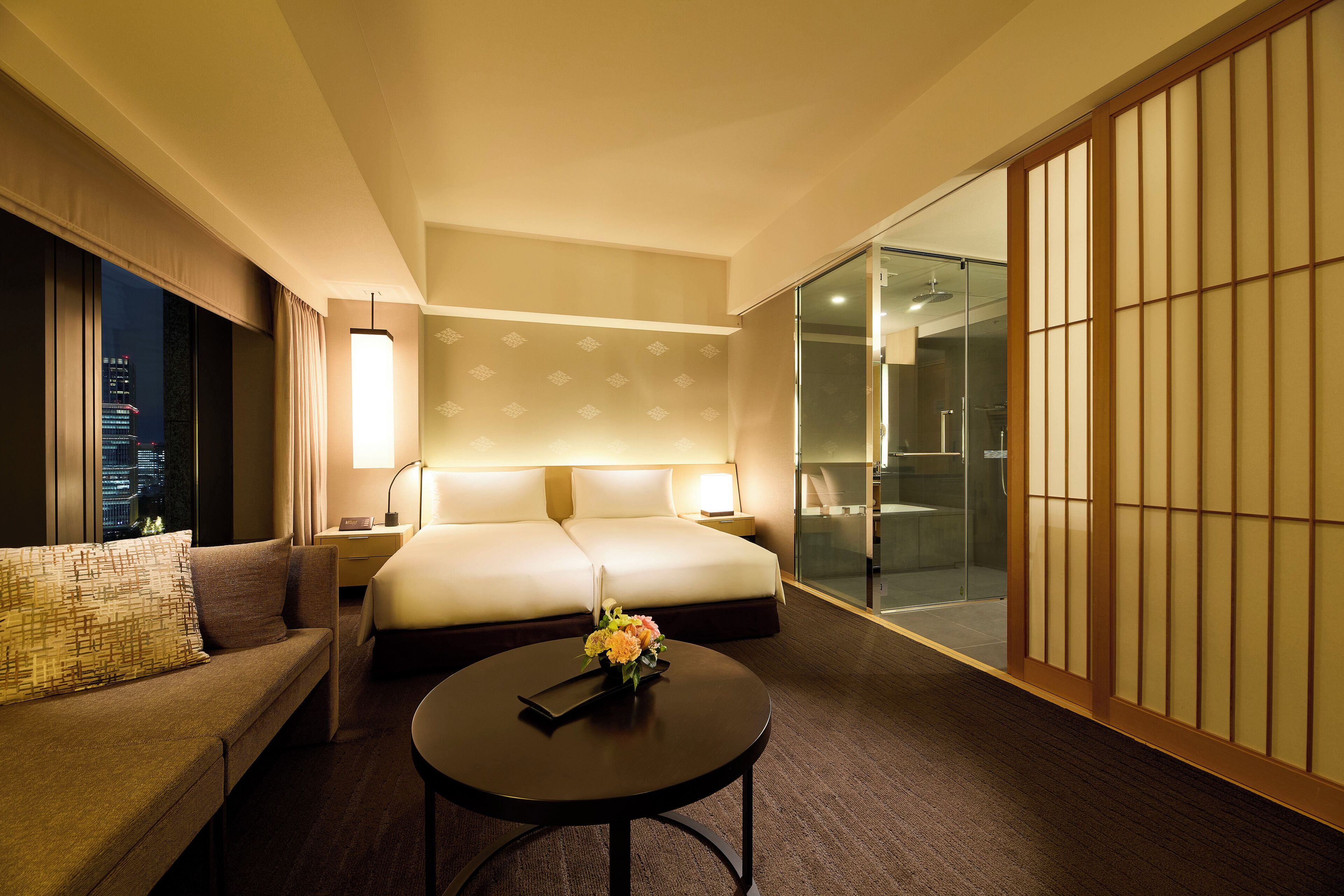 A hotel room with a bed, a sofa, a round coffee table with flowers, a nightstand, a lamp, and a sliding glass door leading to a bathroom.