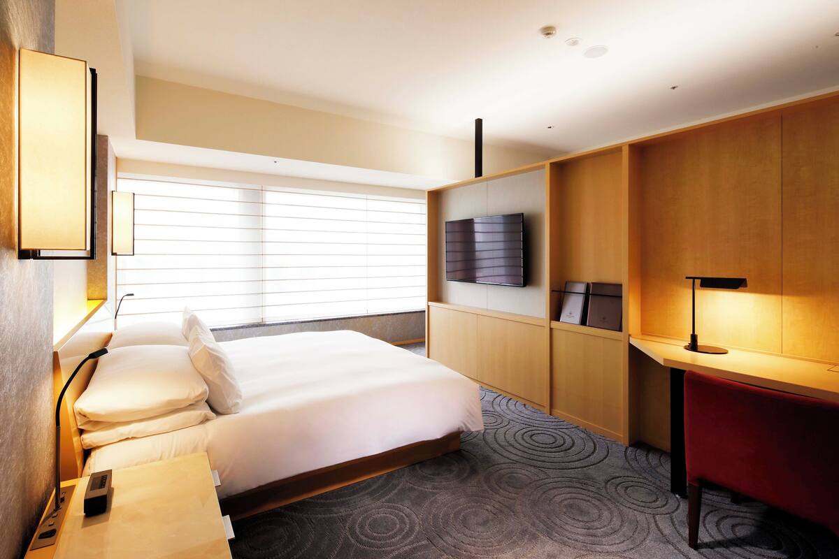 A hotel room with a bed, a desk, a TV, and a window with blinds.