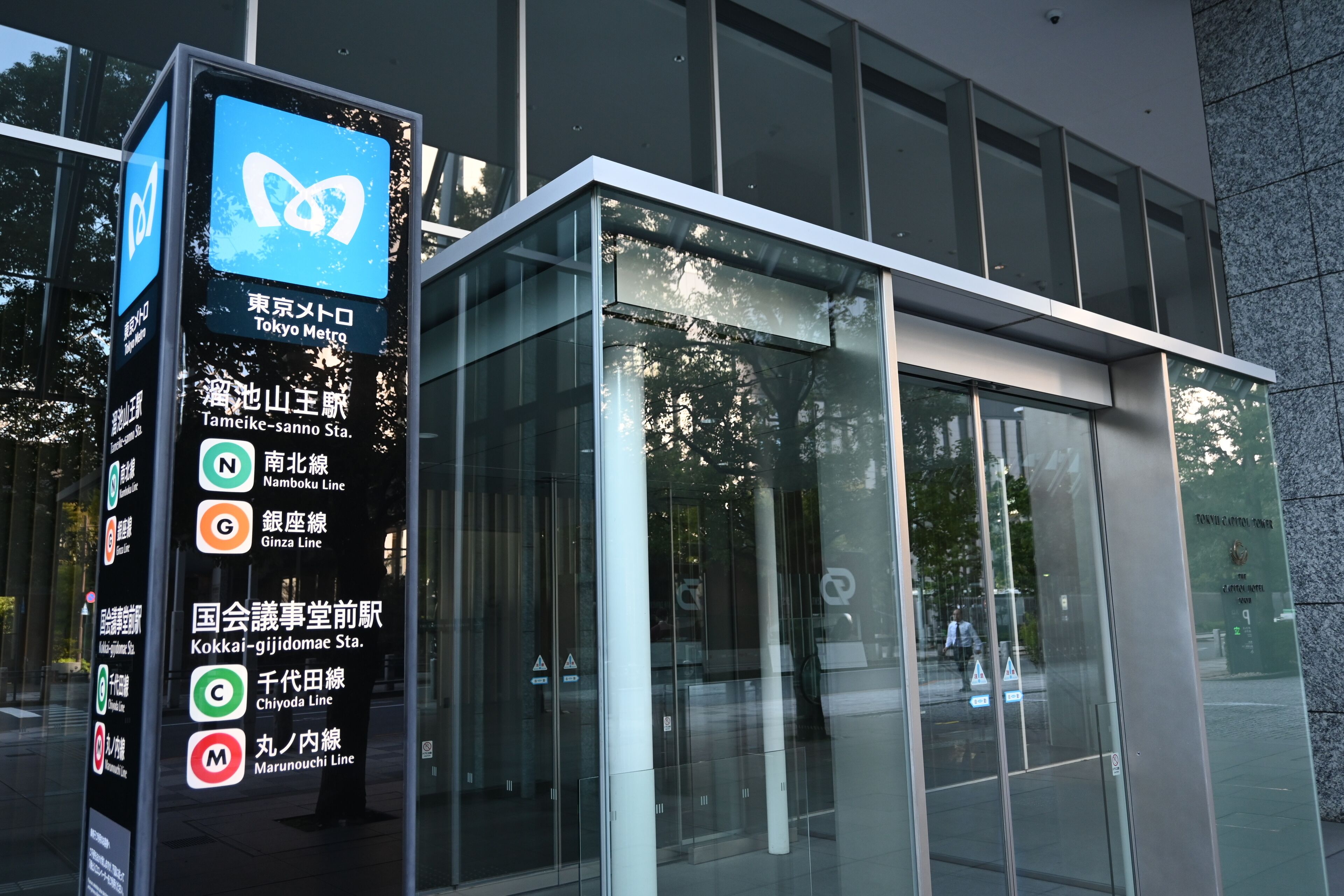 A modern glass building with a digital information board displaying train line names and station names in Japanese.