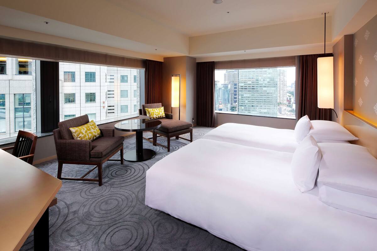 A hotel room with two beds, a sofa, a small table, and large windows offering a city view.