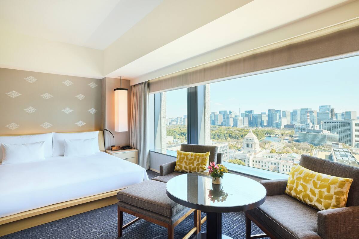 A hotel room with a large bed, a sofa, a small table, and a view of the cityscape.