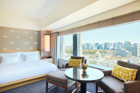 A hotel room with a large bed, a sofa, a small table, and a view of the cityscape.