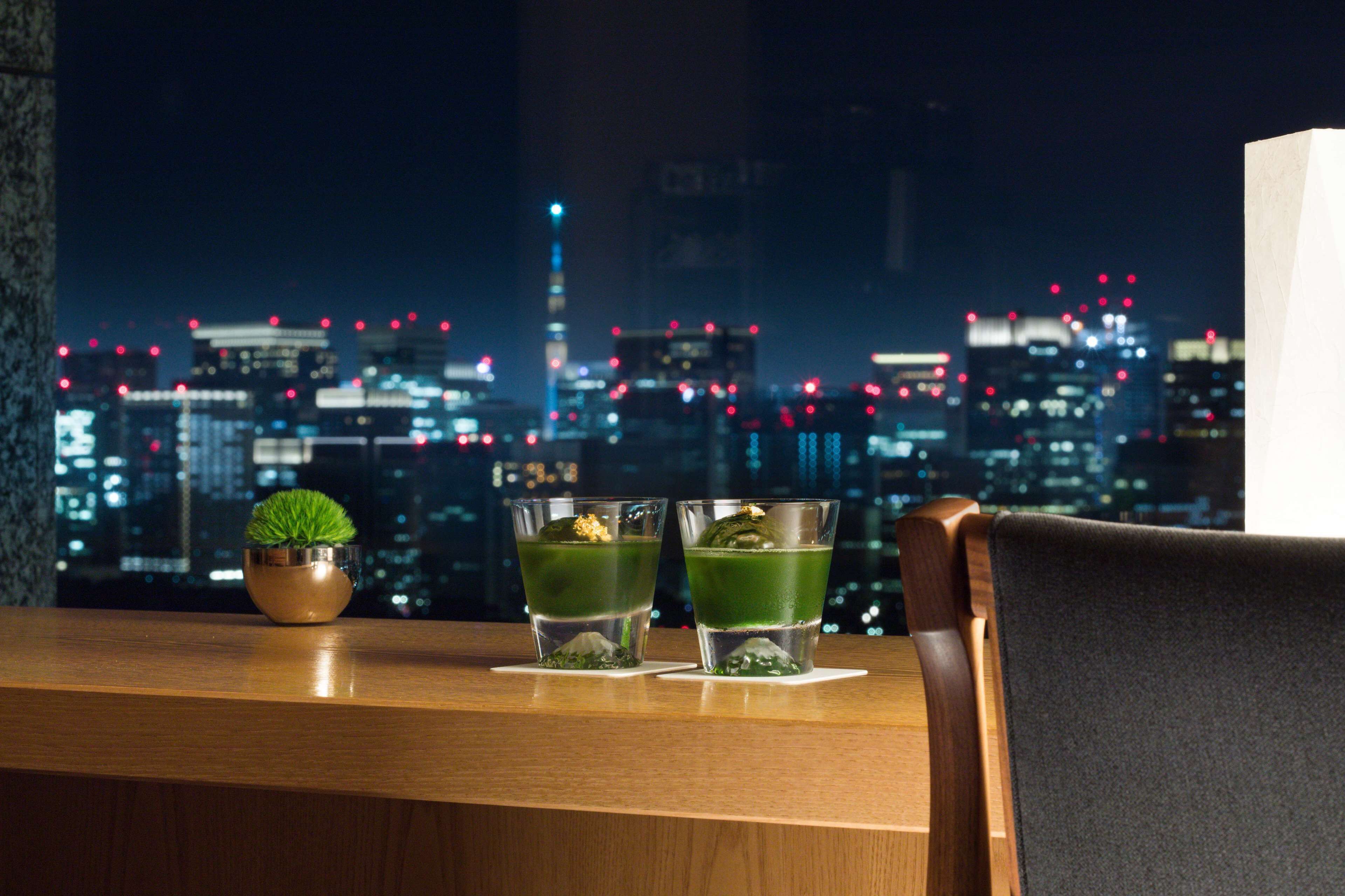 Un bar avec deux verres de cocktail vert, une petite plante en pot et une vue sur la ville de nuit.