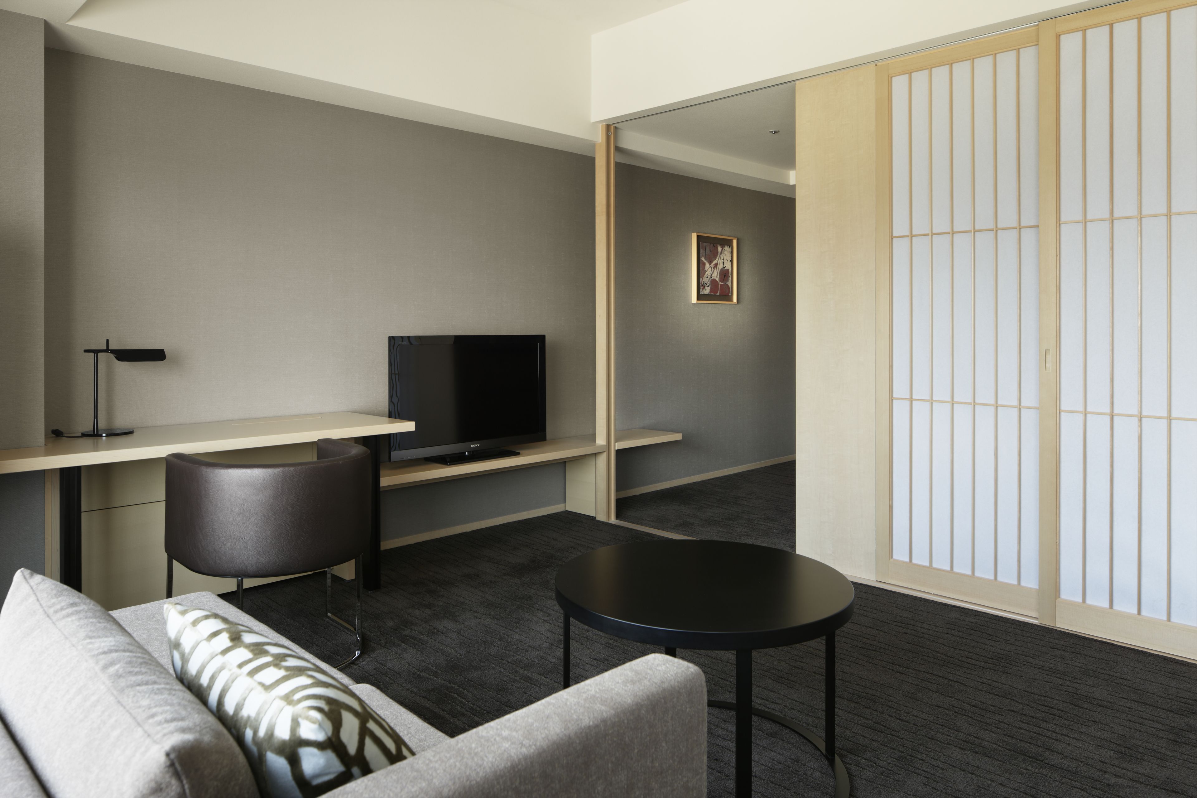 A modern hotel room with a sofa, a small round table, a desk with a television, and a sliding shoji screen.