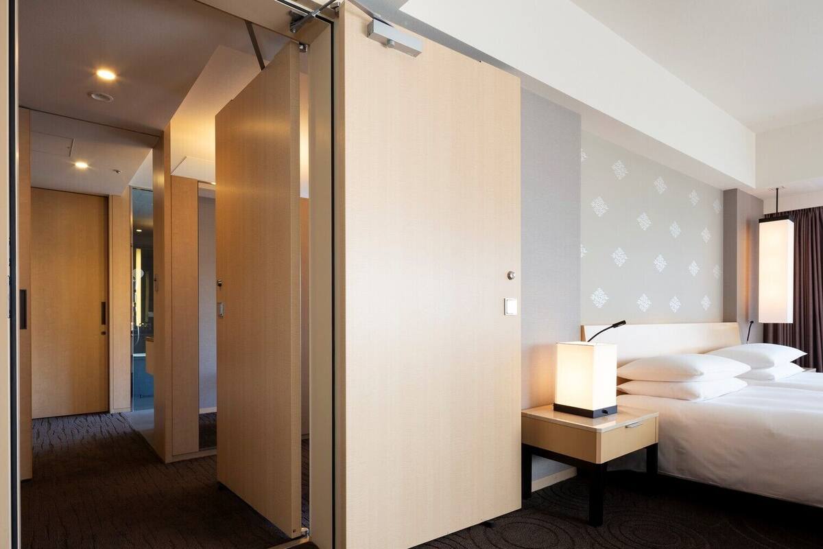 A hotel room with a bed, bedside table, lamp, and a view of the outside.