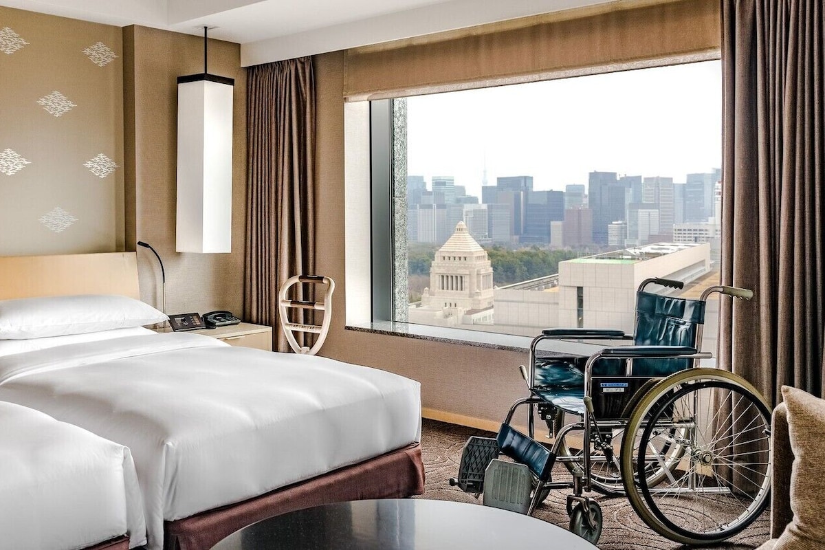 Habitación de hotel con dos camas, una silla de ruedas, vista a la ciudad y un ventanal grande.