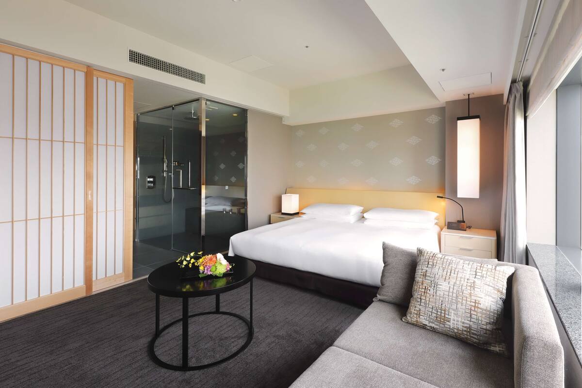 A hotel room with a bed, a sofa, a small table with flowers, a sliding glass door, and a nightstand with a lamp.