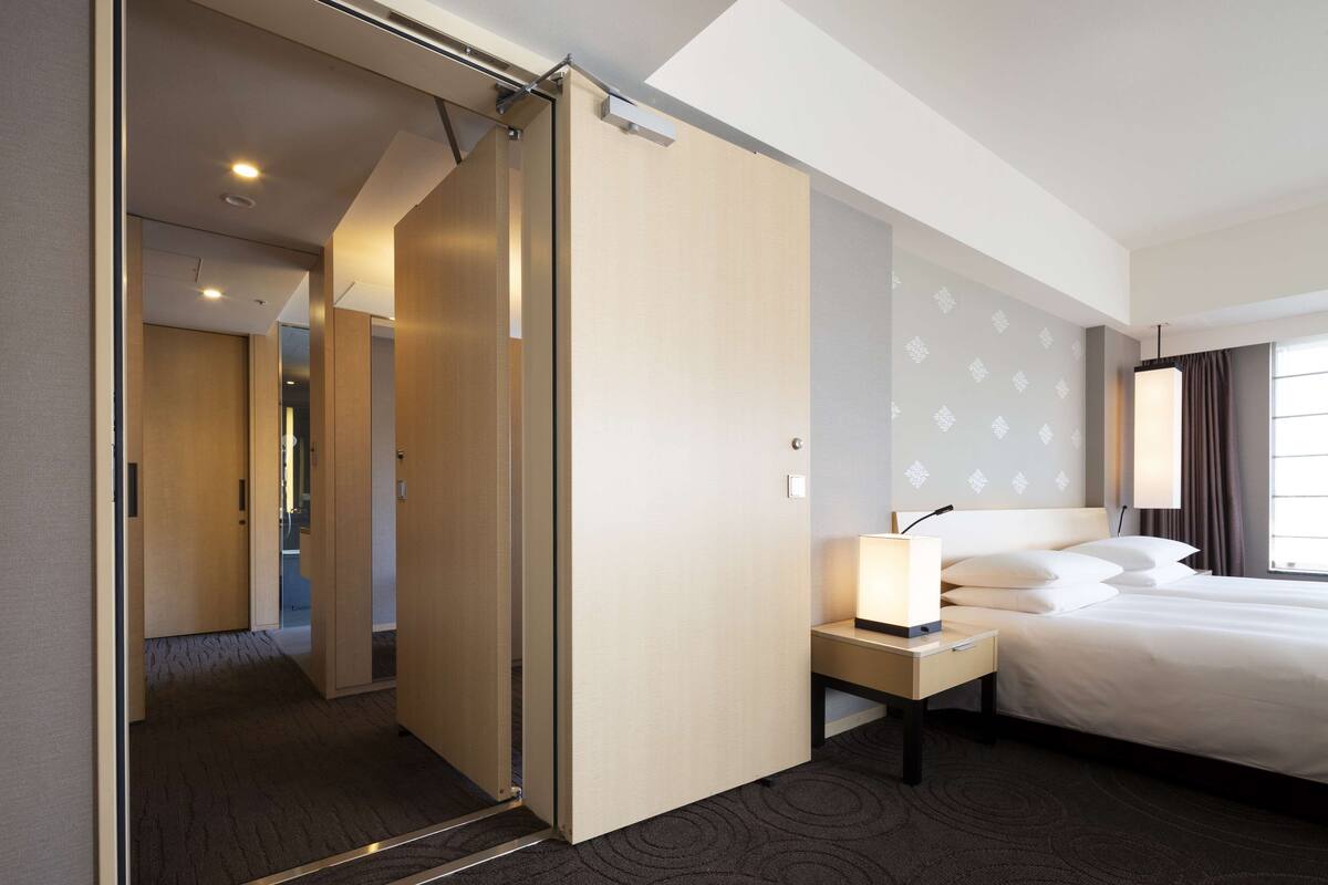 A hotel room with a bed, bedside table, and a view of a hallway.