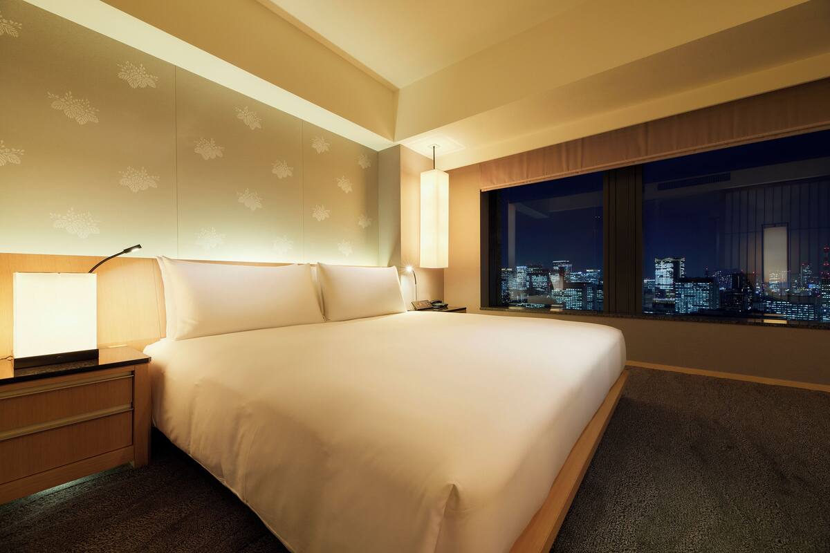 A hotel room with a large bed, a nightstand, a lamp, and a city view through a large window.