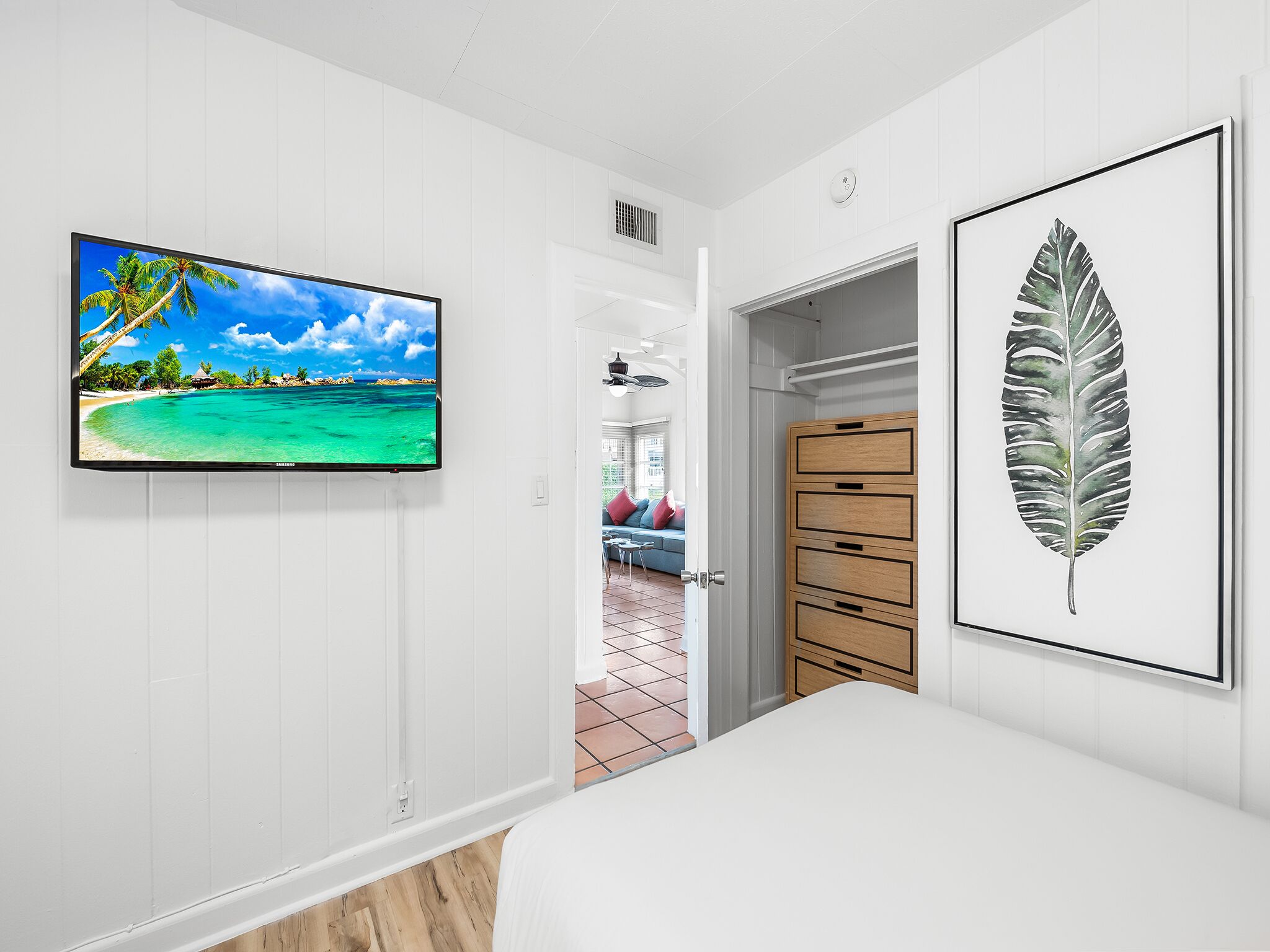 A bedroom with a TV displaying a tropical beach scene, a large leaf artwork, and a wooden wardrobe.