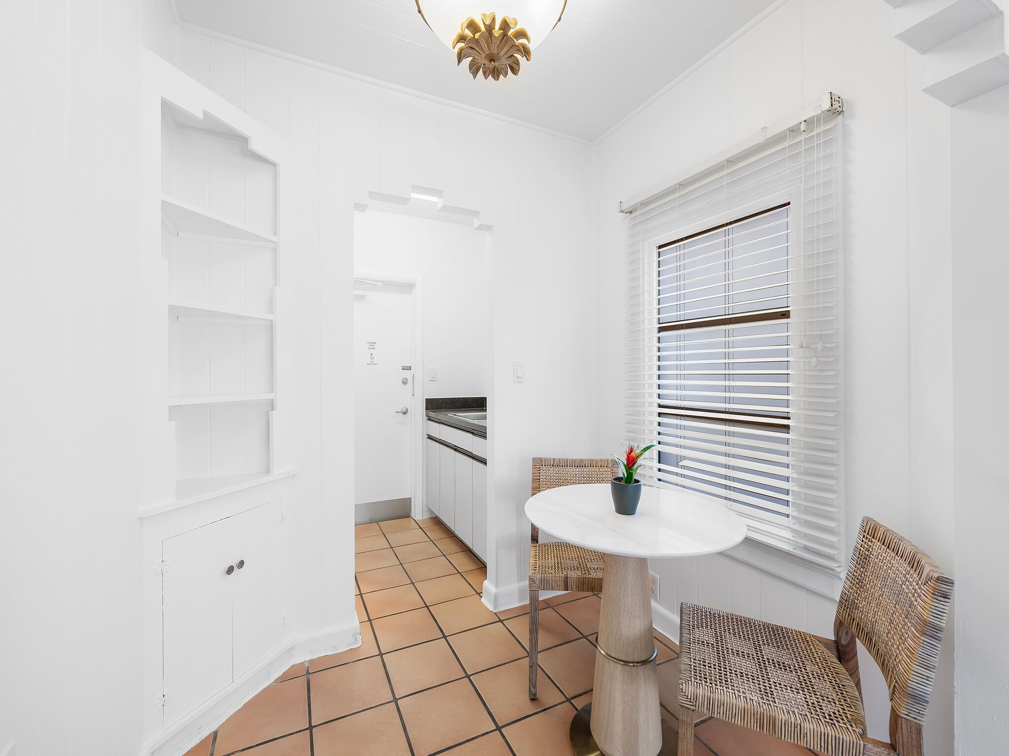 Un interior limpio y blanco con una mesa redonda, sillas de mimbre, una planta en maceta y una ventana con persianas.