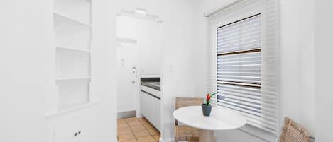 Um interior claro e arejado, com uma mesa redonda e cadeiras de vime, uma planta em vaso e uma janela com persianas.