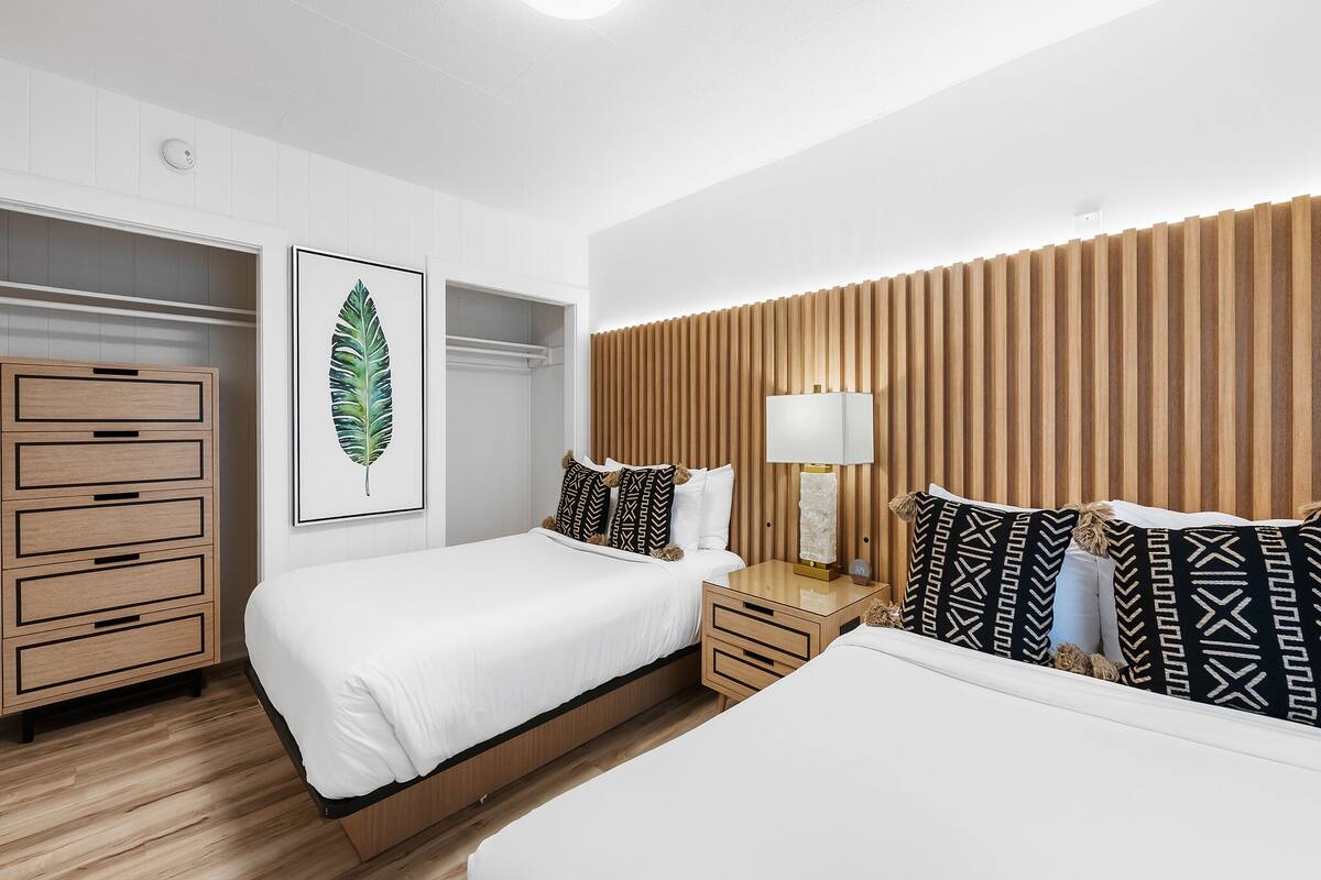 A modern hotel room with two beds, a wooden headboard, a nightstand with a lamp, and a wardrobe with multiple drawers.