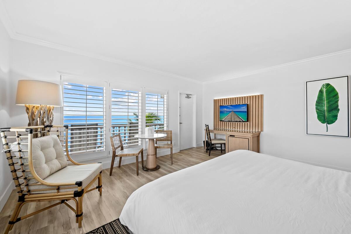 A hotel room with a large bed, a desk with a chair, a round table with chairs, and a view of the ocean through the windows.