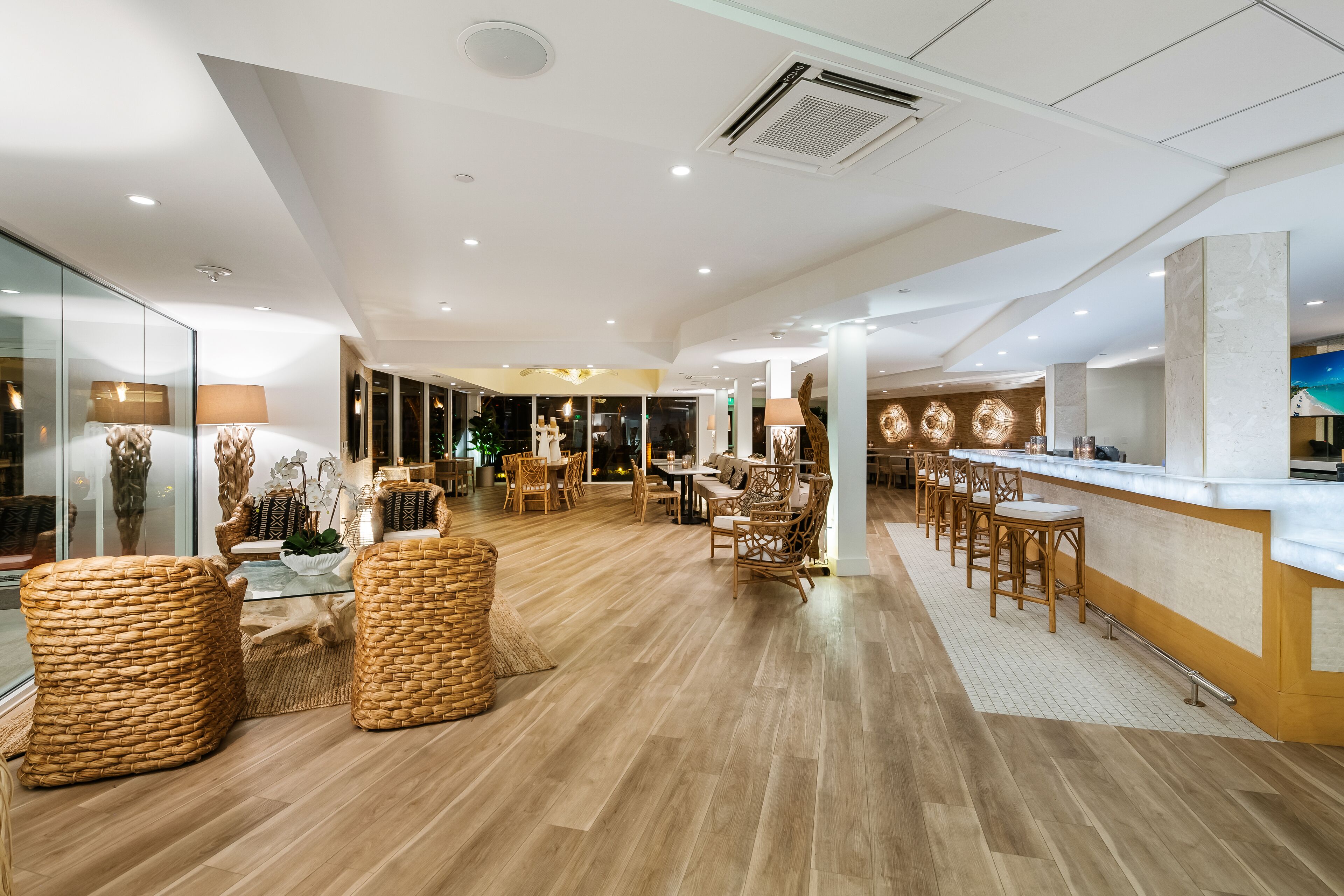 A spacious hotel lobby with a wooden floor, wicker furniture, a bar area, and a view of the cityscape through large windows.