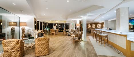 Un amplio vestíbulo de hotel con suelo de madera, mobiliario de mimbre, una zona de bar y vistas a la ciudad a través de amplias ventanas.