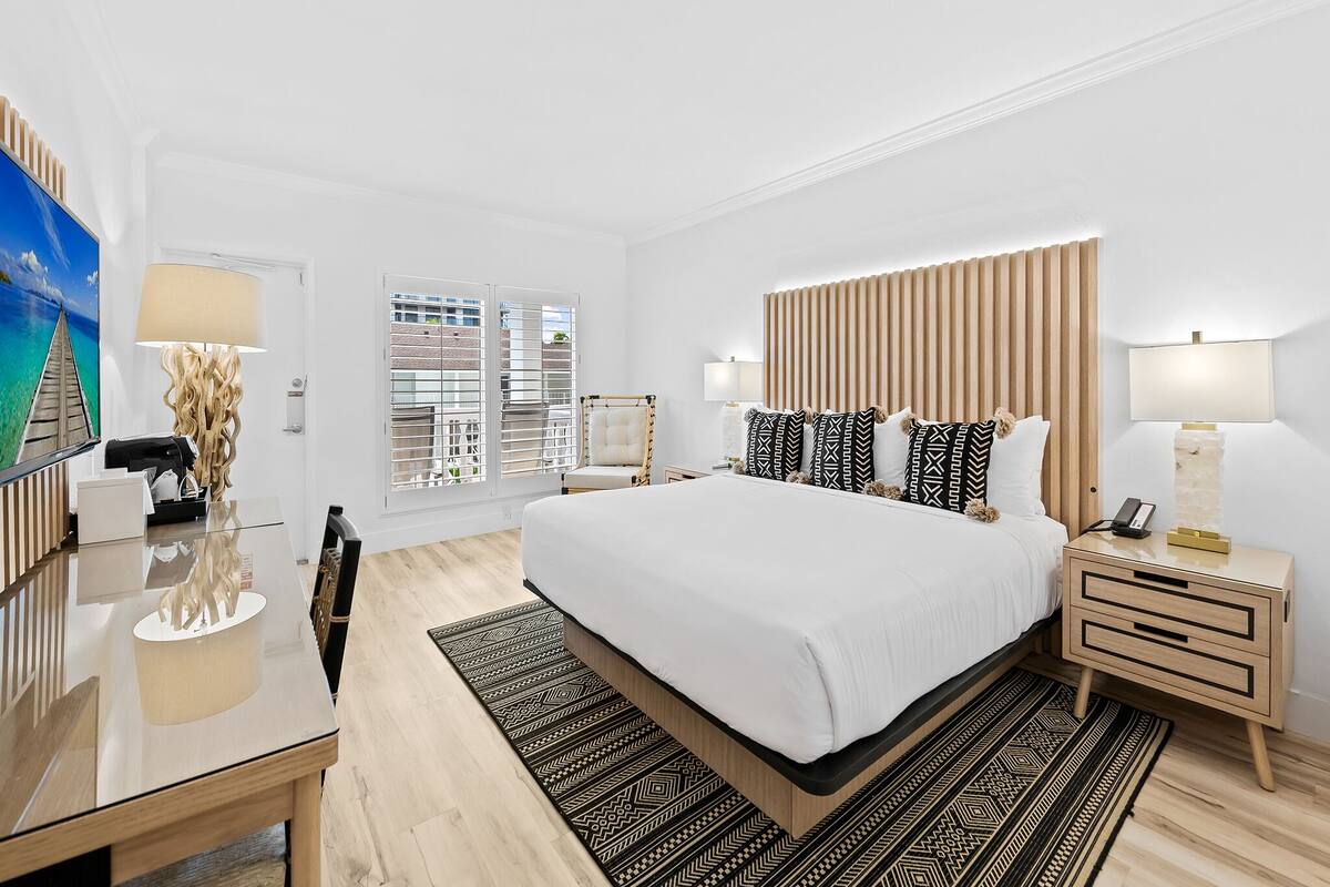 A modern hotel room with a large bed, bedside tables, a desk, and a view through the window.