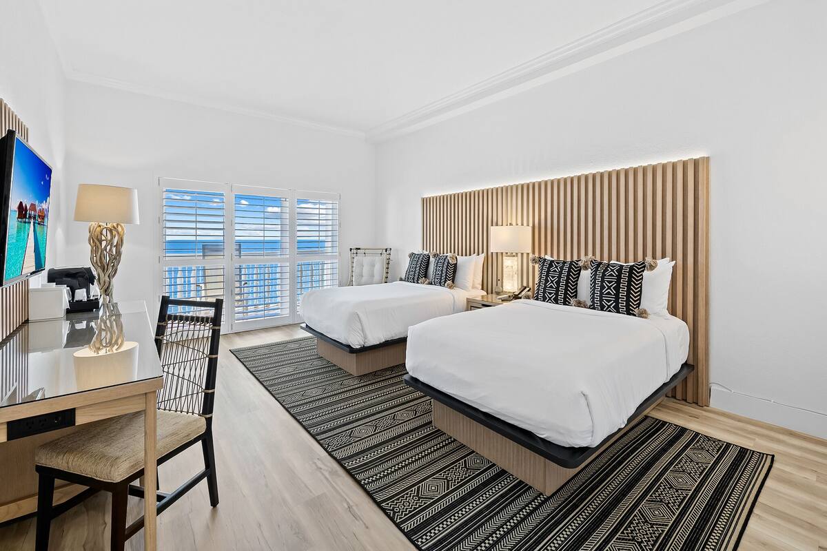 A hotel room with two beds, a desk, a chair, and a large window with blinds.