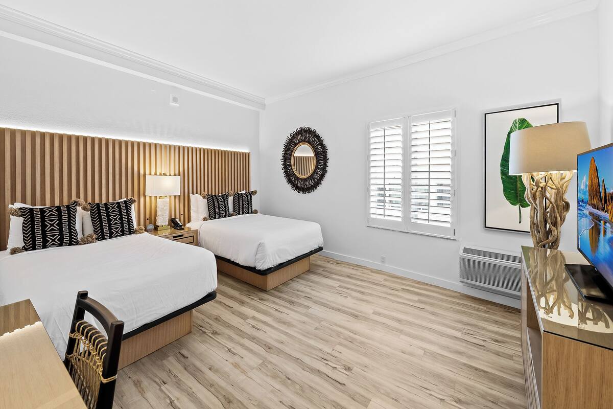 A hotel room with two beds, a wooden headboard, a bedside table with a lamp, a wall-mounted mirror, a framed picture, and a radiator.