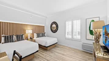Habitación de hotel con dos camas, cabecero de madera, mesita de noche con lámpara, espejo colgado en la pared, cuadro enmarco y radiador.