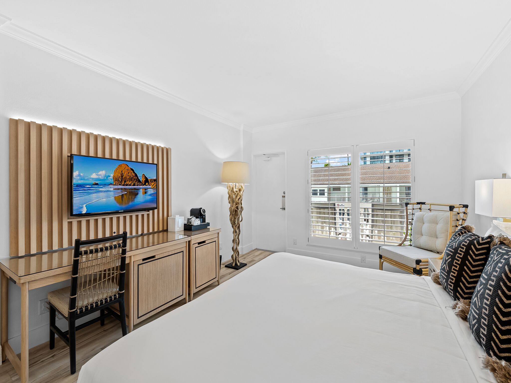 A hotel room with a bed, a desk with a chair, a TV mounted on a wooden panel, a lamp, and a window with a view of buildings.