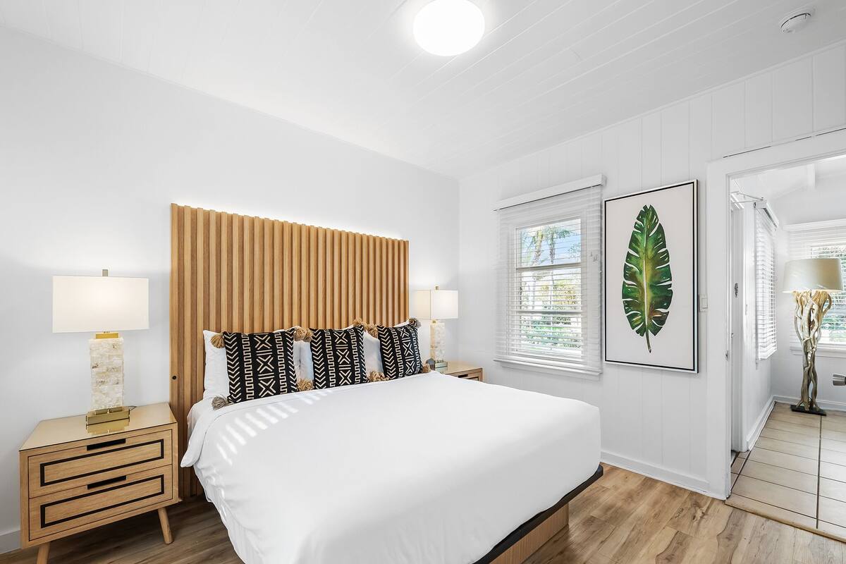 A bedroom with a wooden headboard, a bed with white bedding, two bedside tables with lamps, and a large leaf artwork on the wall.
