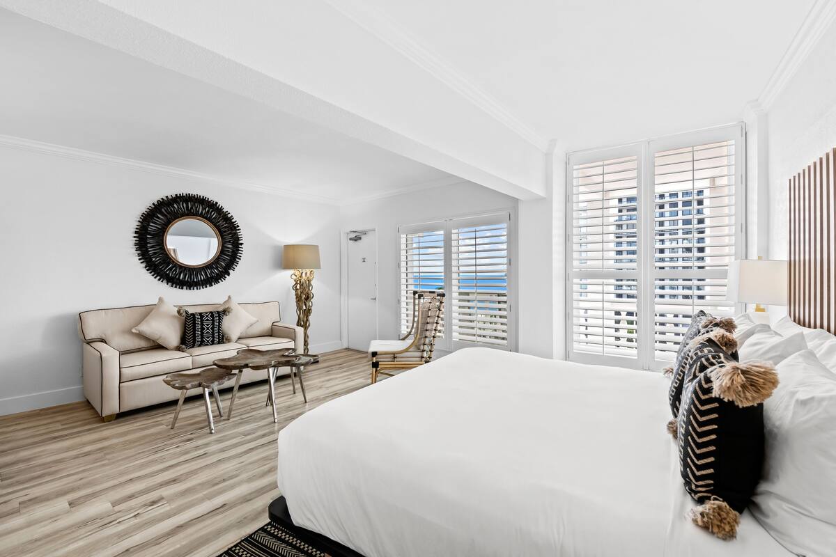 A hotel room with a large bed, a sofa, a chair, a coffee table, and a view of the city through the windows.