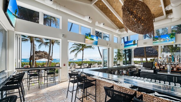 A spacious outdoor bar area with multiple seating options, a bar counter, and large windows offering views of palm trees and the ocean. - Beachcomber Resort & Club (Pompano Beach)