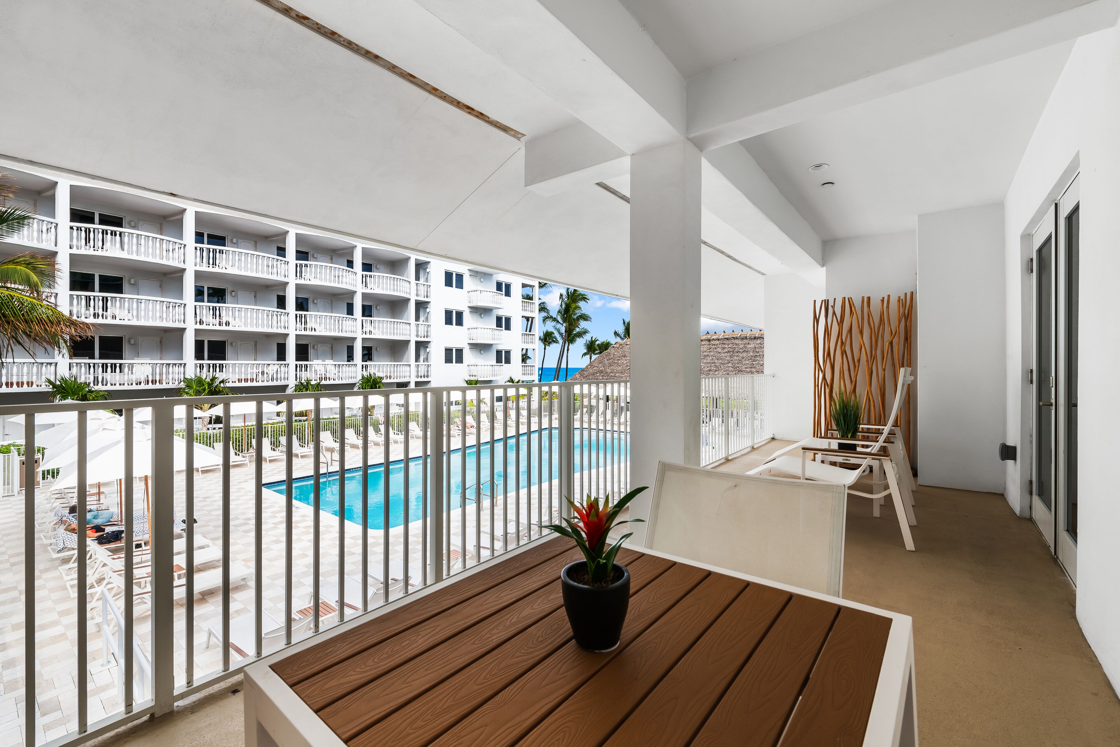 A balcony with a wooden table and a potted plant, overlooking a swimming pool and a building with balconies.