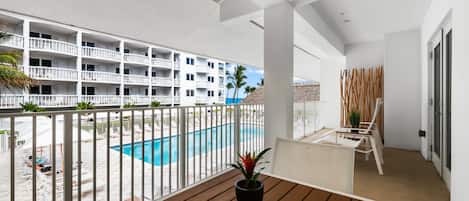 A balcony with a wooden table and a potted plant, overlooking a swimming pool and a building with balconies.