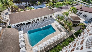 Um resort com piscina rodeada por espreguiçadeiras e guarda-sóis, área de estar coberta e palmeiras.