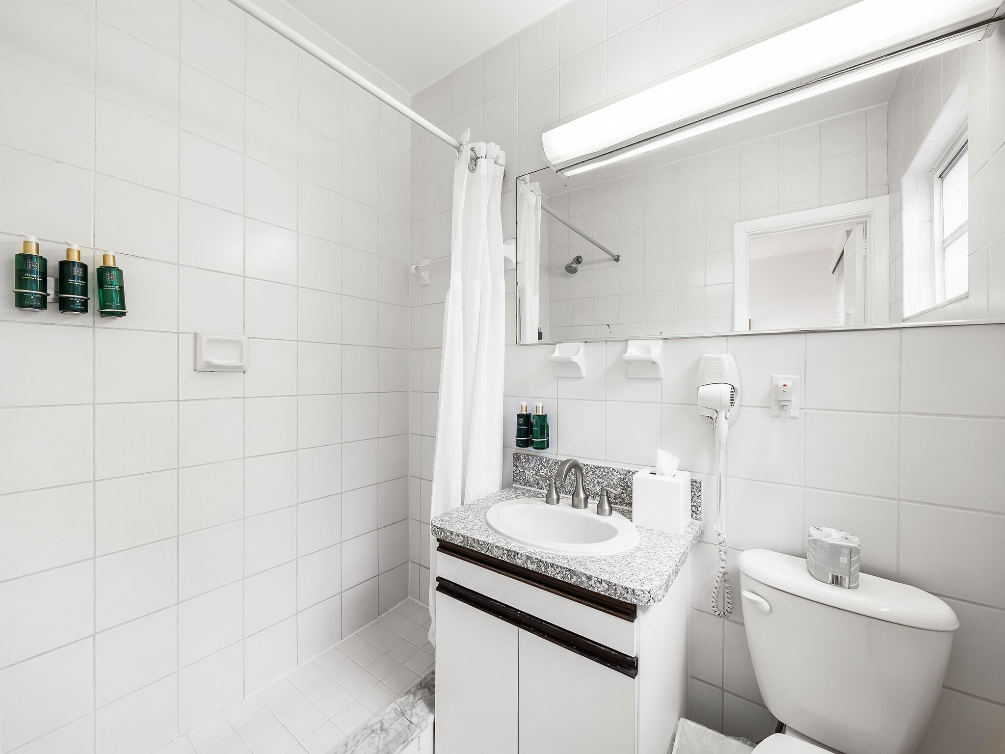 A modern bathroom with white tiles, a shower, a sink with a granite countertop, and a toilet.