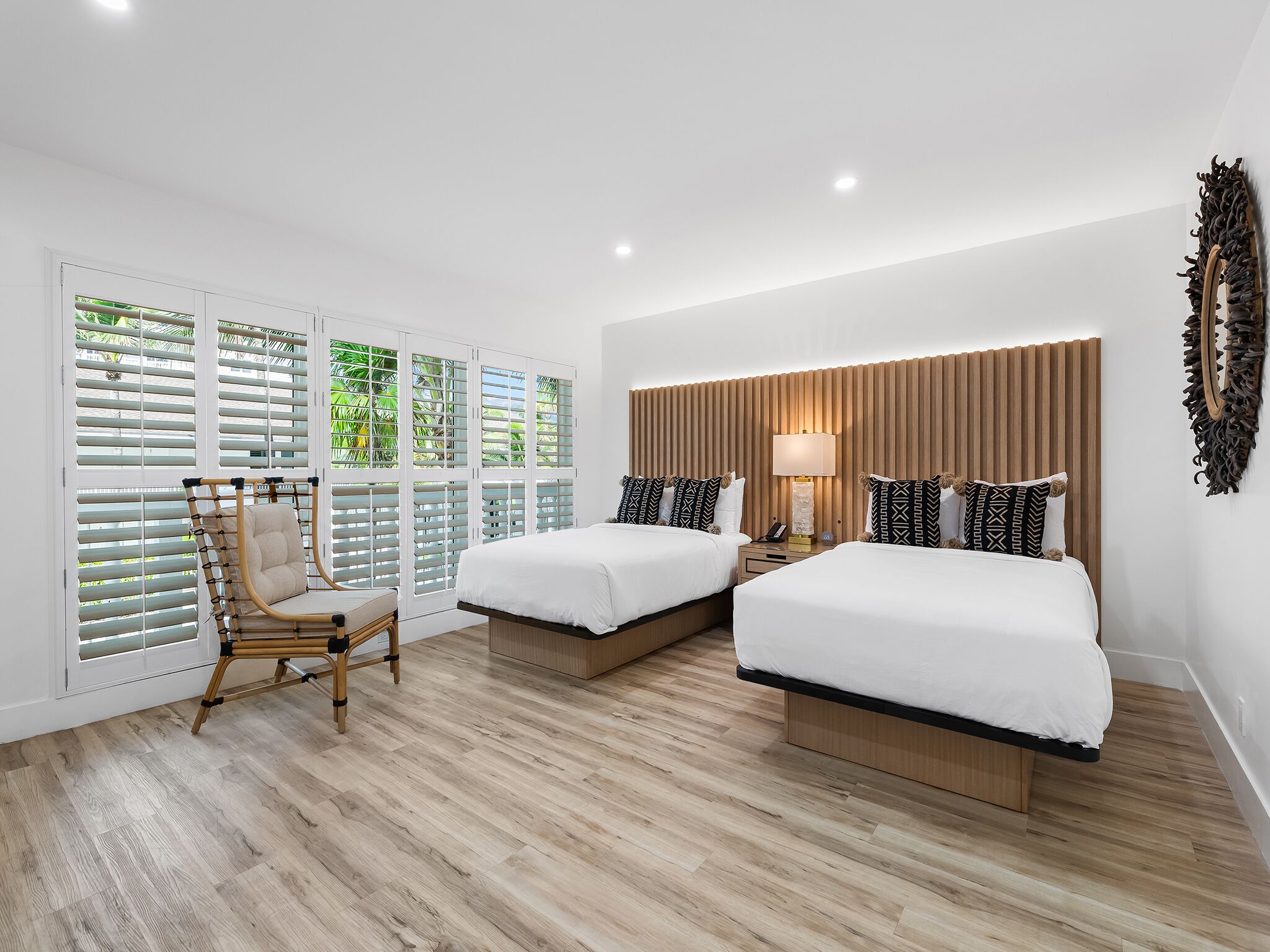 A hotel room with two beds, a wooden floor, a chair, and a view through large windows with shutters.