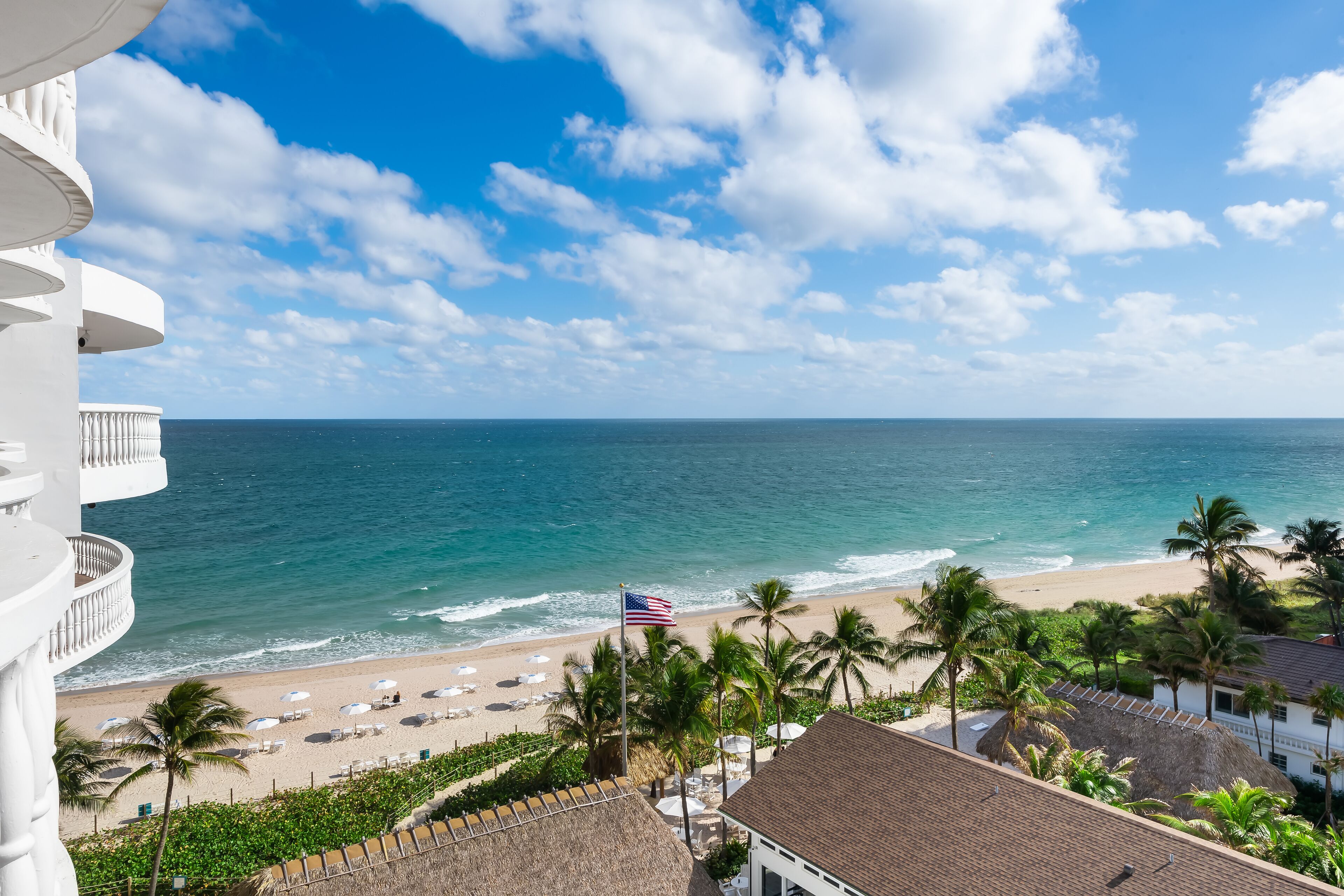 A beachfront hotel with a balcony overlooking the ocean, a flag, and palm trees.