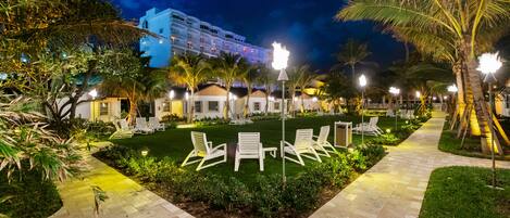 A well-lit outdoor area with a paved walkway, lounge chairs, and palm trees.