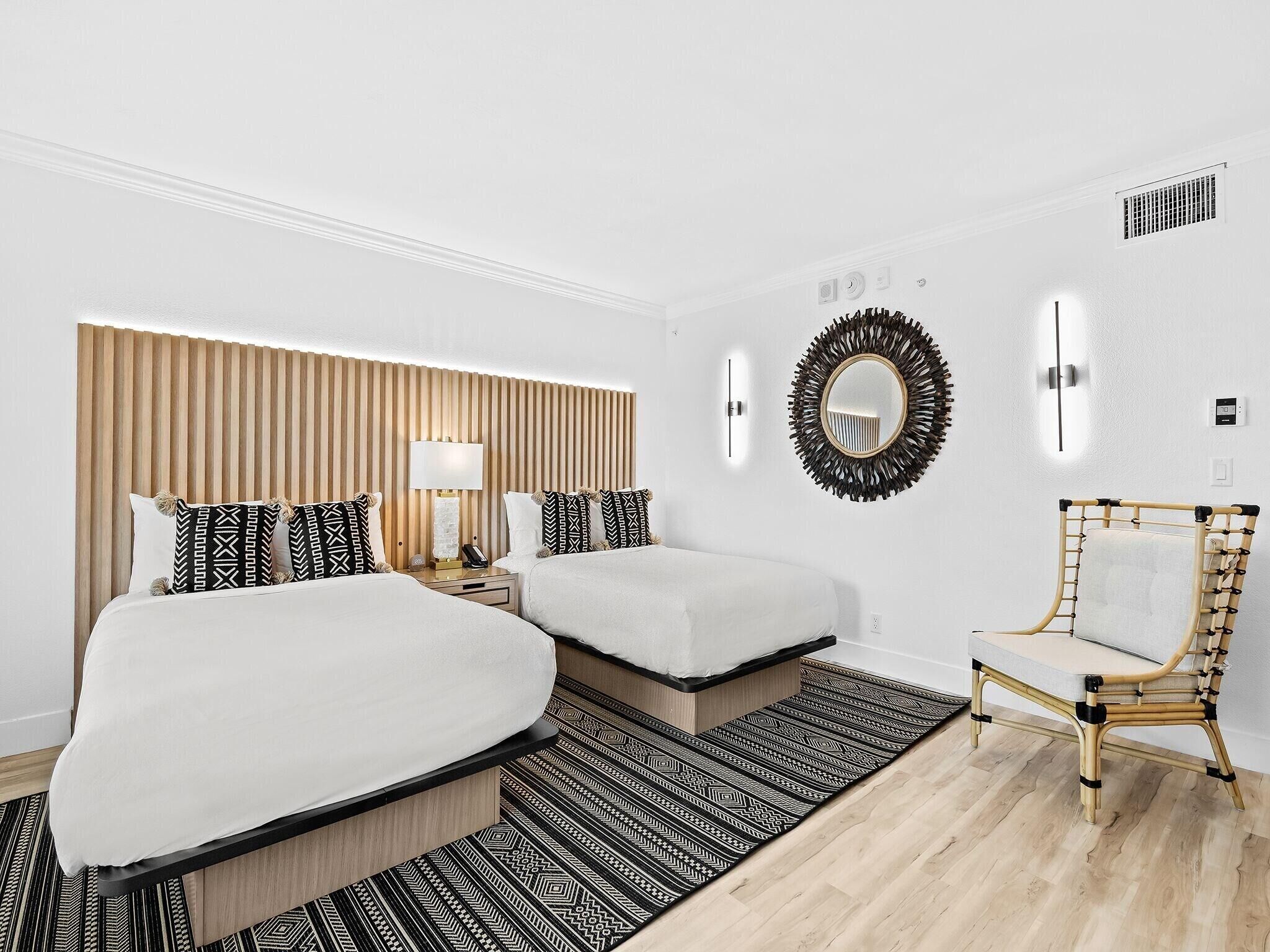 Habitación de hotel con dos camas, una mesita de noche con lámpara, un espejo, una silla y una alfombra estampada.
