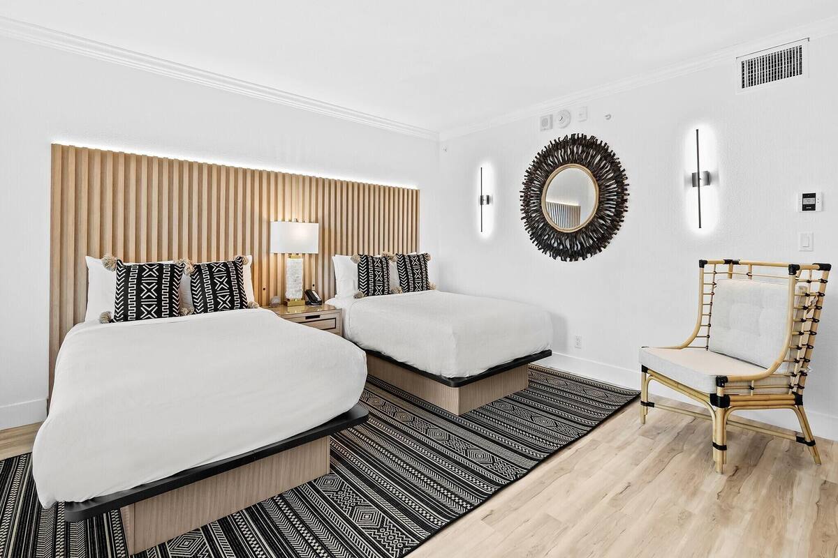 A hotel room with two beds, a bedside table with a lamp, a mirror, a chair, and a patterned rug.