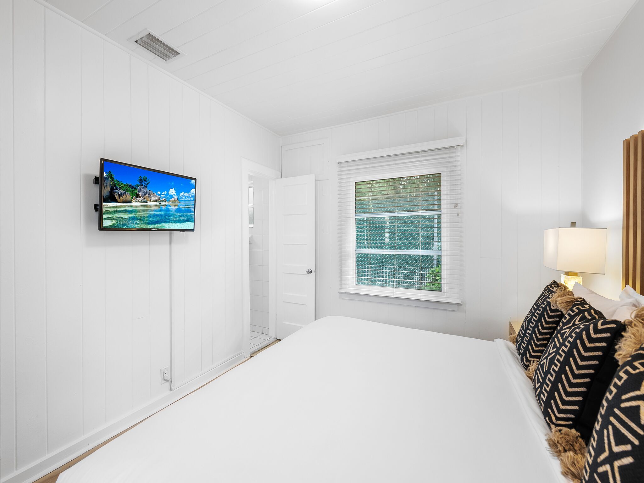 A hotel room with a bed, a TV mounted on the wall, a window with blinds, and a bedside lamp.