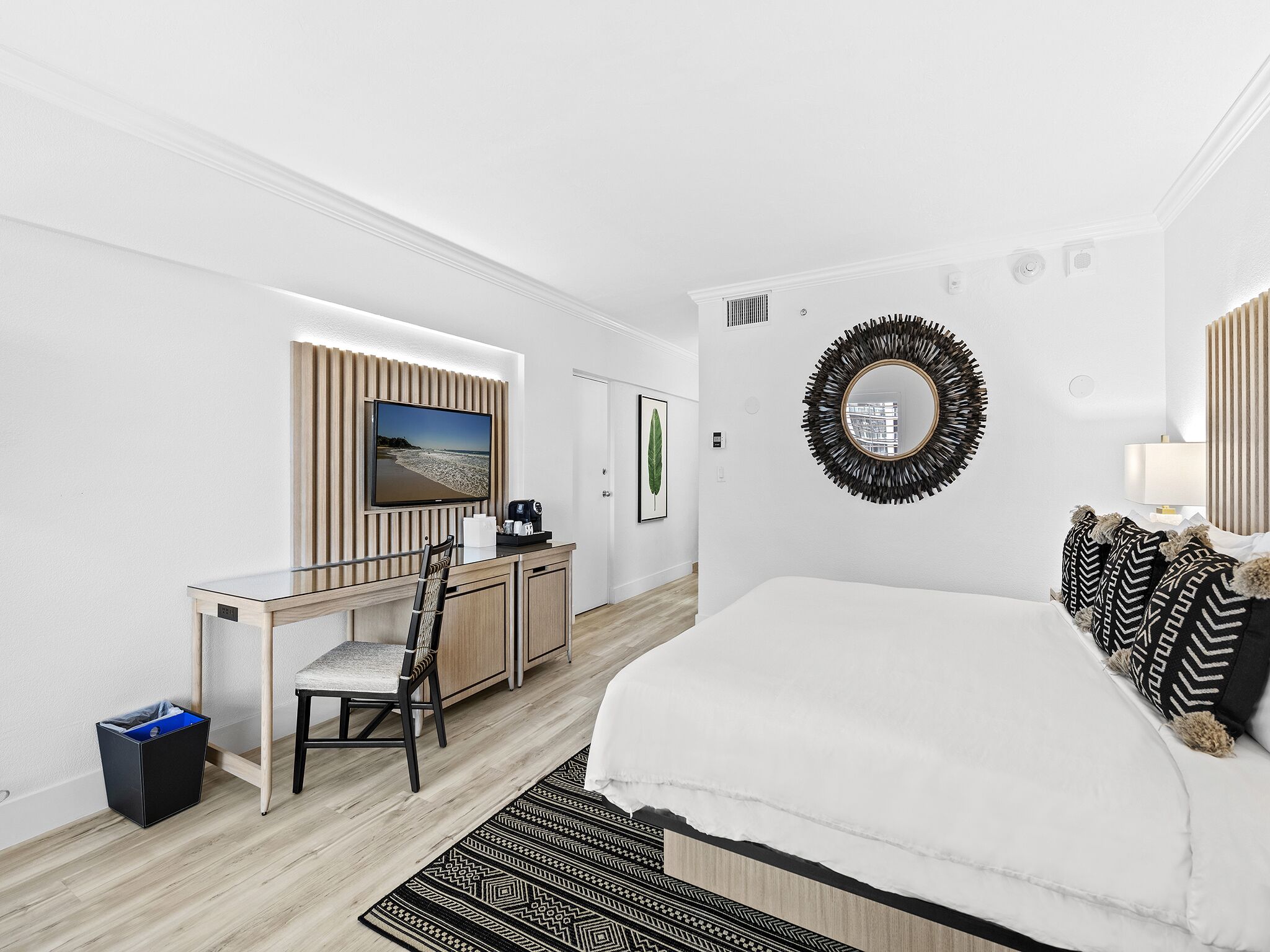 Una habitación de hotel moderna con cama, escritorio con silla, televisor, espejo y un cesto de basura.