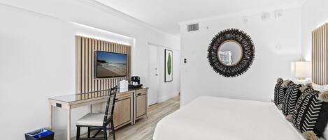 Una habitación de hotel moderna con cama, escritorio con silla, televisor, espejo y un cesto de basura.