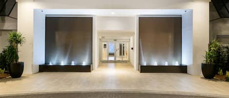 A modern hotel entrance with a light wood floor, large potted plants, and a glass door leading to the interior.