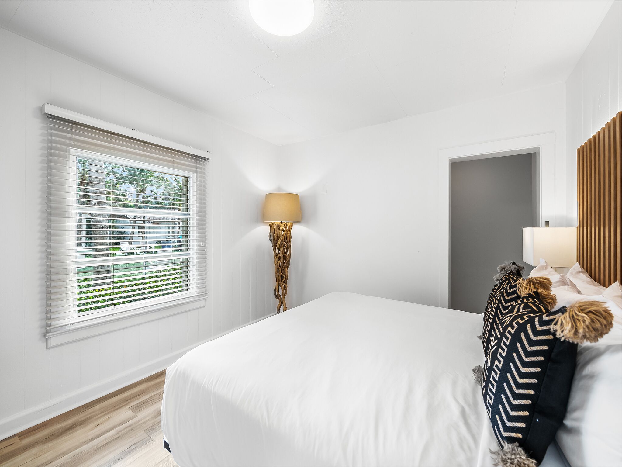A bedroom with a large bed, a bedside lamp, a window with blinds, and a door.